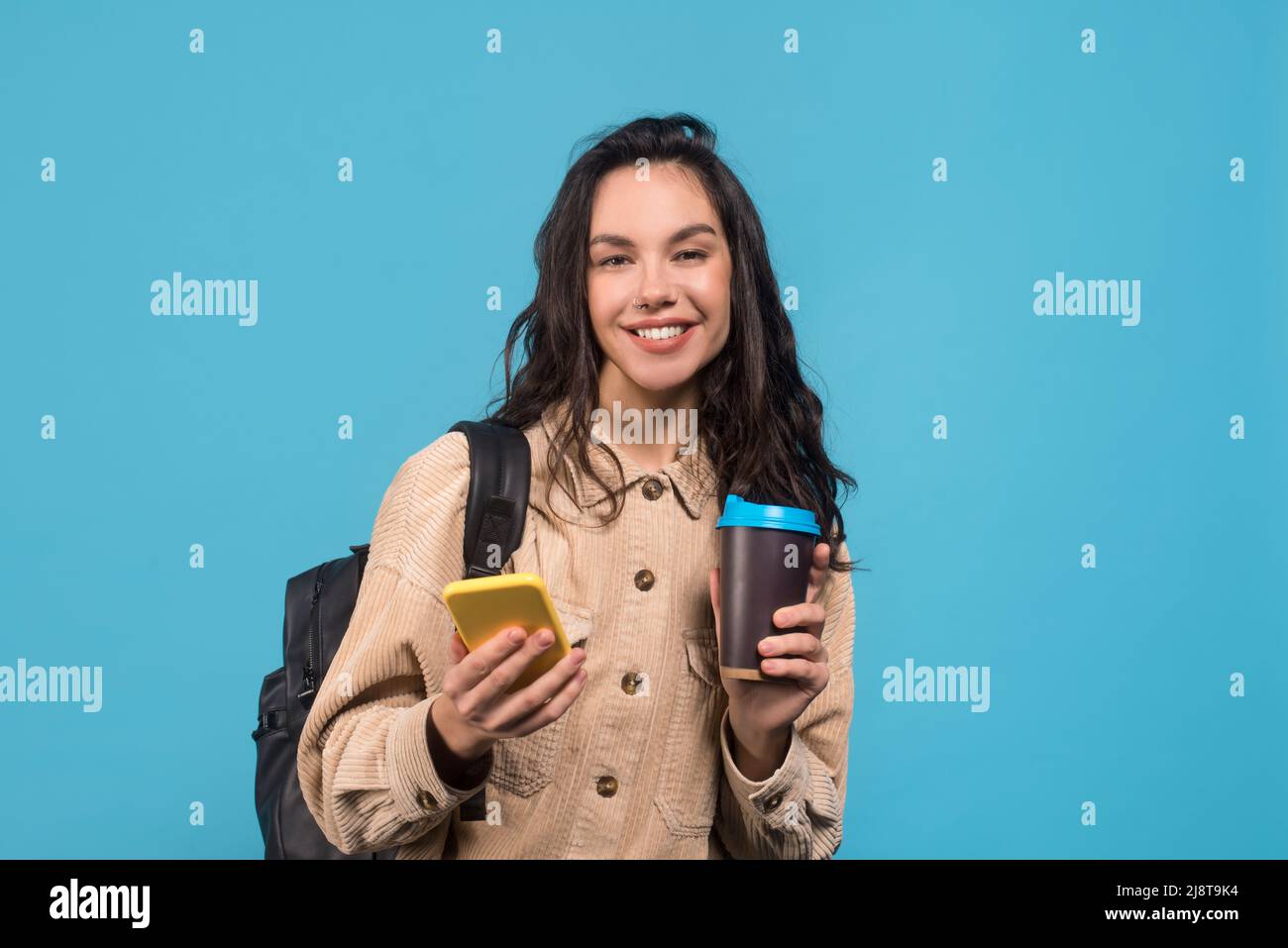 Jeune femme brunette européenne souriante avec sac à dos, tasse de café et smartphone Banque D'Images