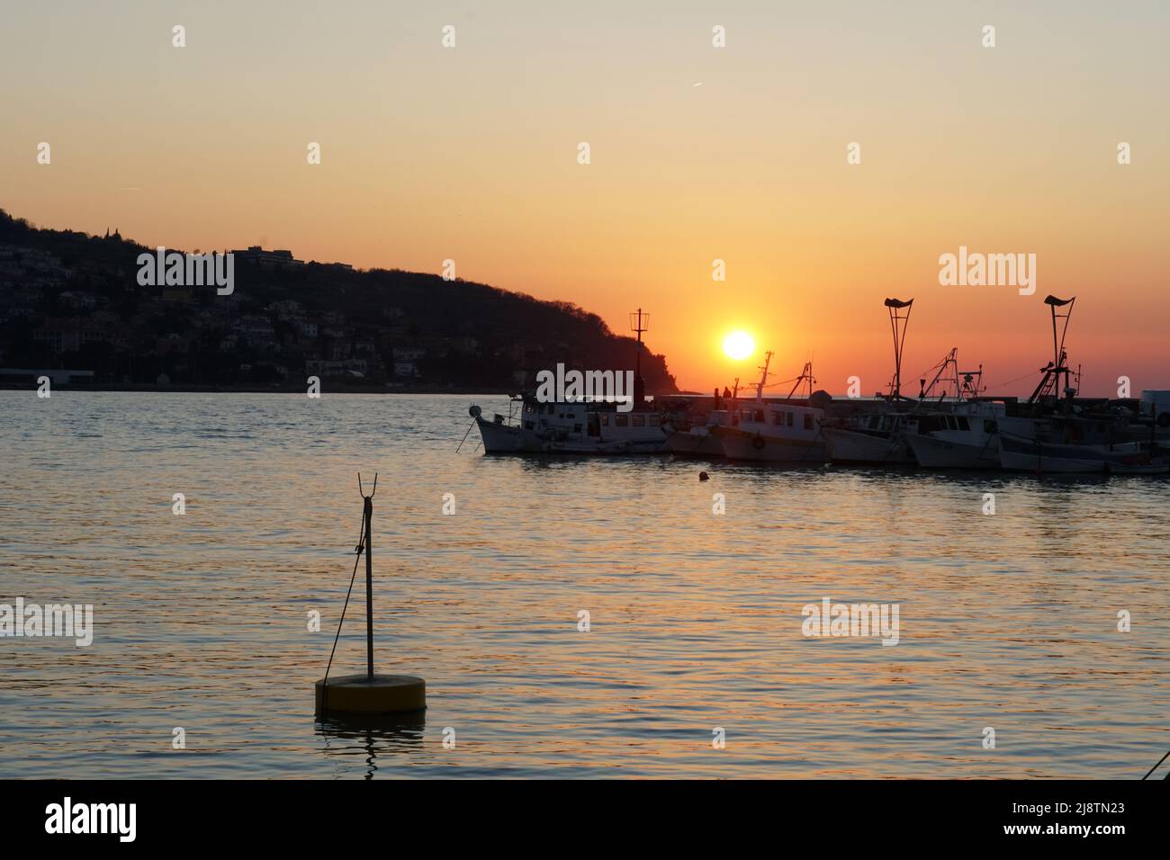 Coucher de soleil dans le port de pêche de Koper, Slovénie. En arrière-plan sont les bateaux et la côte sous le ciel orange spectaculaire. Bouée flottante jaune isolée au premier plan. Banque D'Images