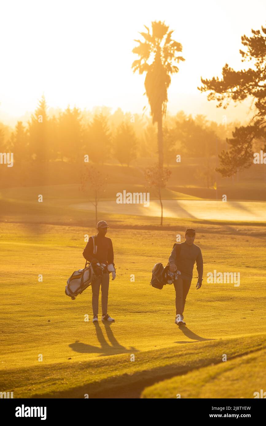 Pleine longueur de jeunes amis multiraciaux avec des sacs marchant contre le ciel clair au parcours de golf Banque D'Images