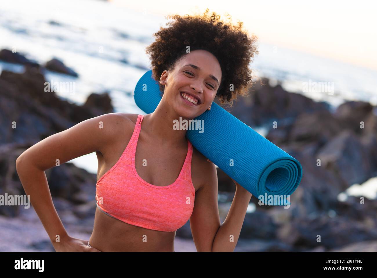 Portrait de jeune femme afro-américaine joyeuse avec tapis de yoga roulé sur l'épaule à la plage Banque D'Images
