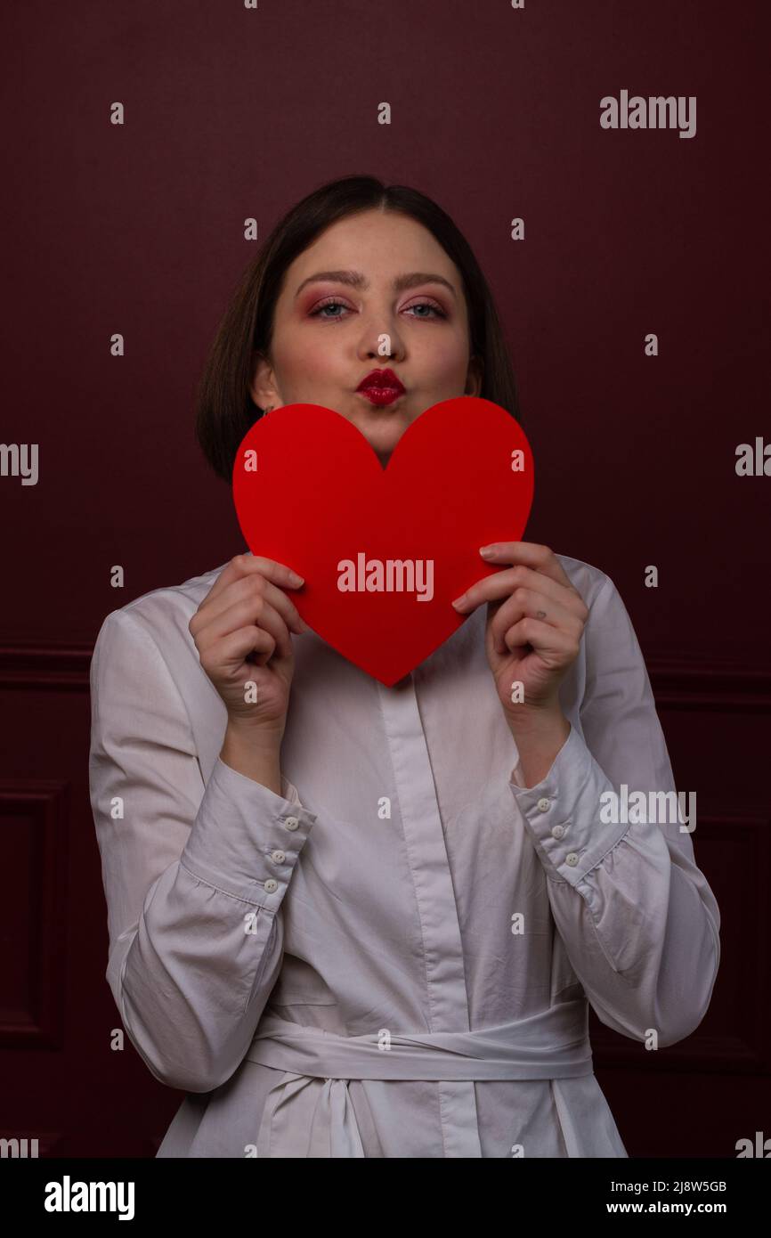 Femme à poil court sur fond rouge tenant le cœur rouge devant son menton avec des lèvres formant un baiser Banque D'Images