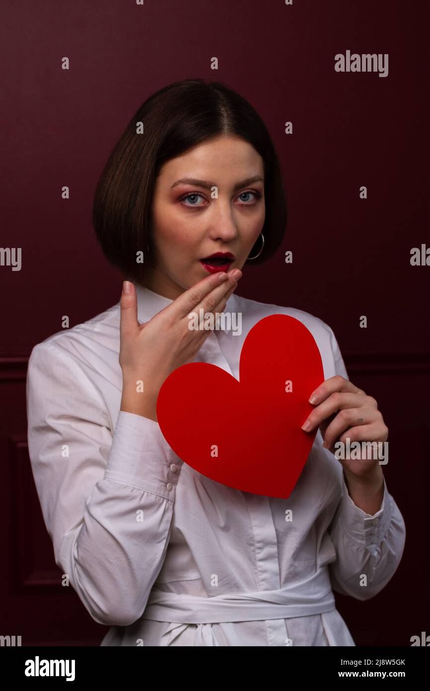 Surpris femme à poil court sur fond rouge tenant la forme de coeur rouge devant elle avec Banque D'Images