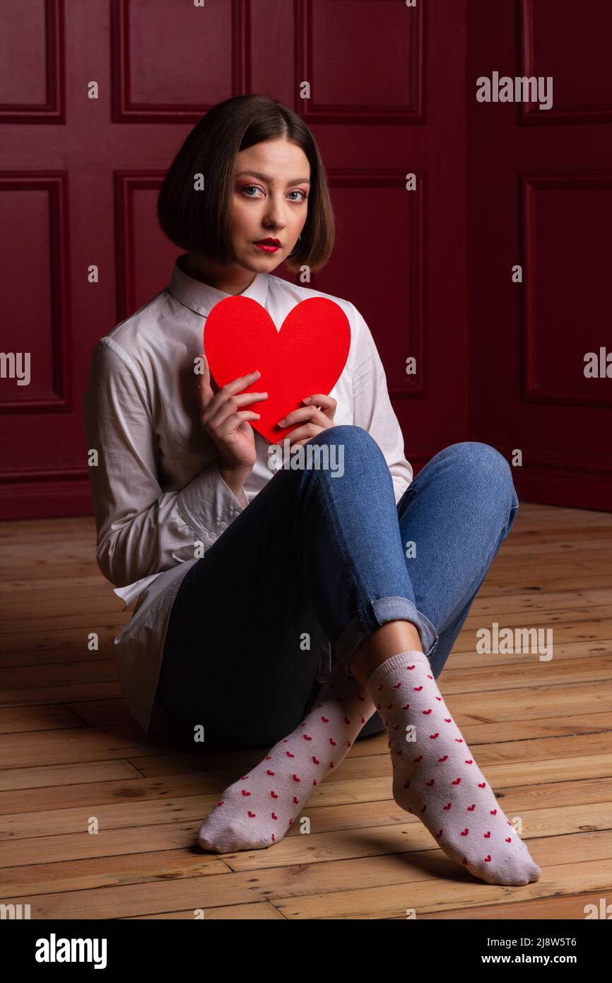 Femme aux cheveux courts regardant dans l'appareil photo assise sur des jambes en bois croisées et arrière-plan rouge tenant la forme de coeur rouge devant sa poitrine Banque D'Images