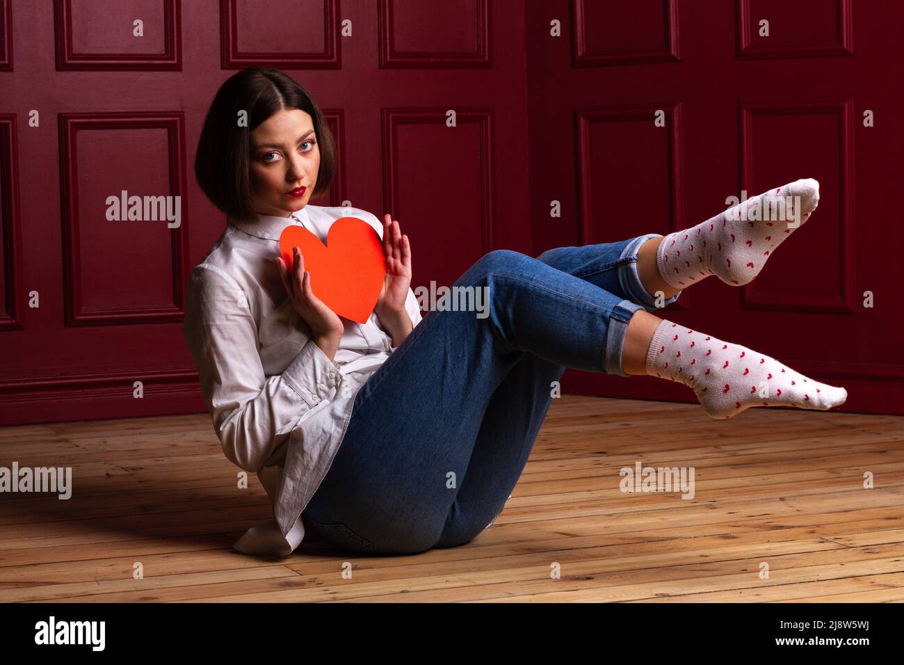 Femme aux cheveux courts assise sur des jambes en bois dans l'air et sur fond rouge tenant le coeur rouge devant sa poitrine Banque D'Images