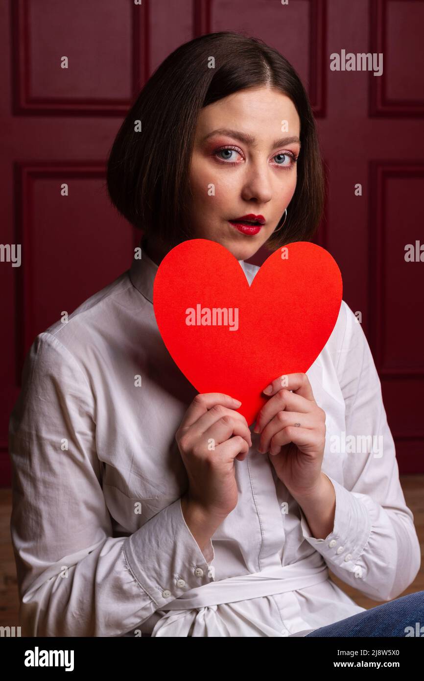 Gros plan d'une femme surprise à poil court sur fond rouge tenant la forme du cœur rouge devant elle Banque D'Images