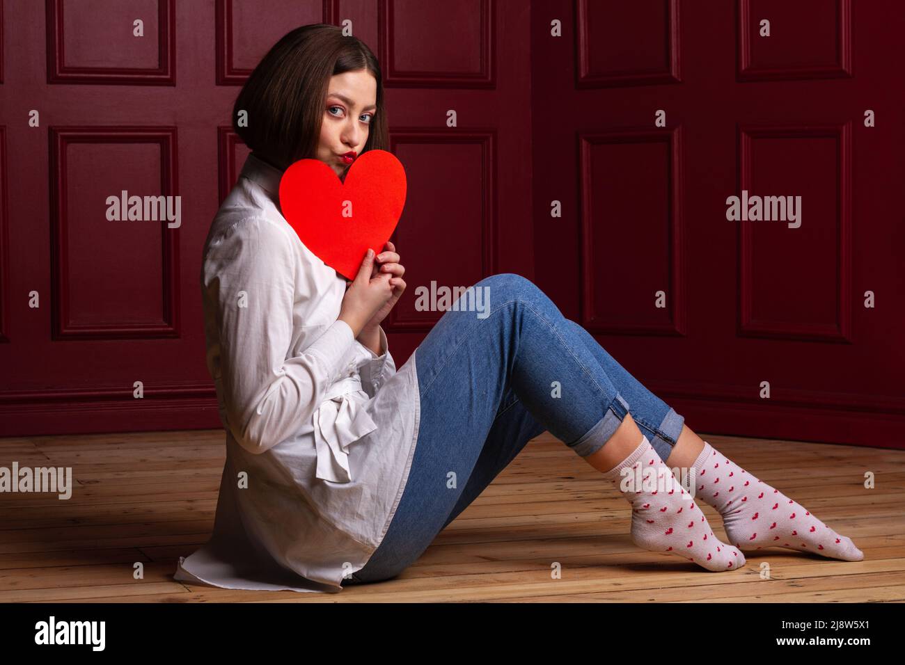 Femme à poil court avec des lèvres formant un baiser assis à côté de l'appareil photo sur un sol en bois et un fond rouge tenant la forme du cœur rouge devant son menton Banque D'Images
