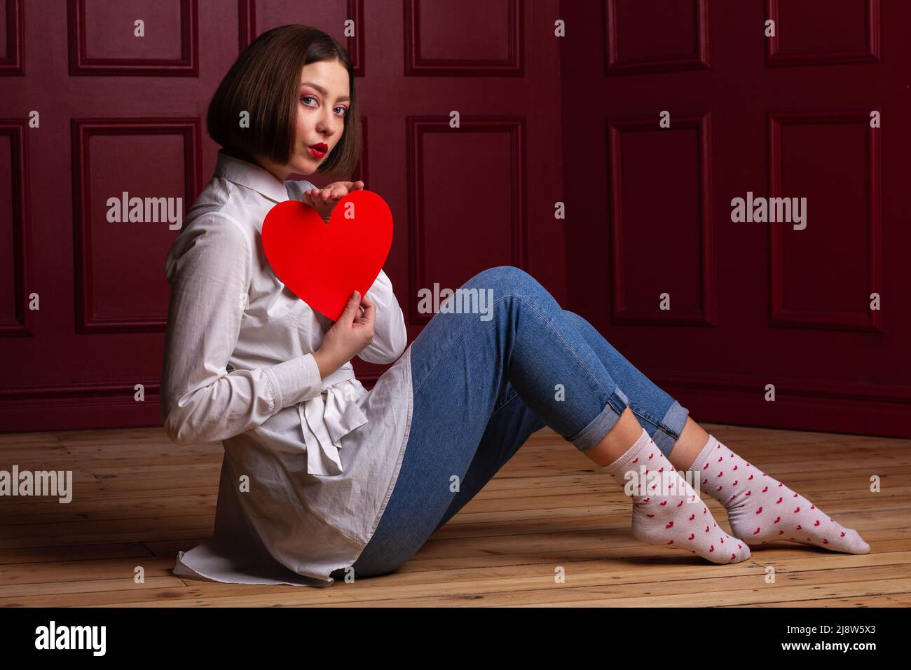 Femme soufflante de baiser assis à côté de l'appareil photo sur parquet et arrière-plan rouge tenant la forme de coeur rouge devant sa poitrine Banque D'Images