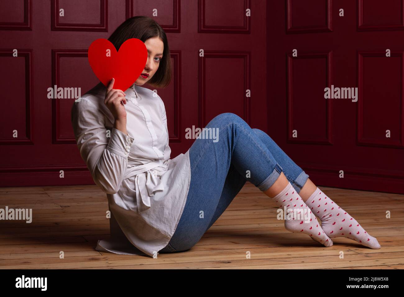 Femme aux cheveux courts assise à côté de l'appareil photo sur un sol en bois et un arrière-plan rouge tenant le coeur rouge devant elle et se cachant le visage derrière elle Banque D'Images