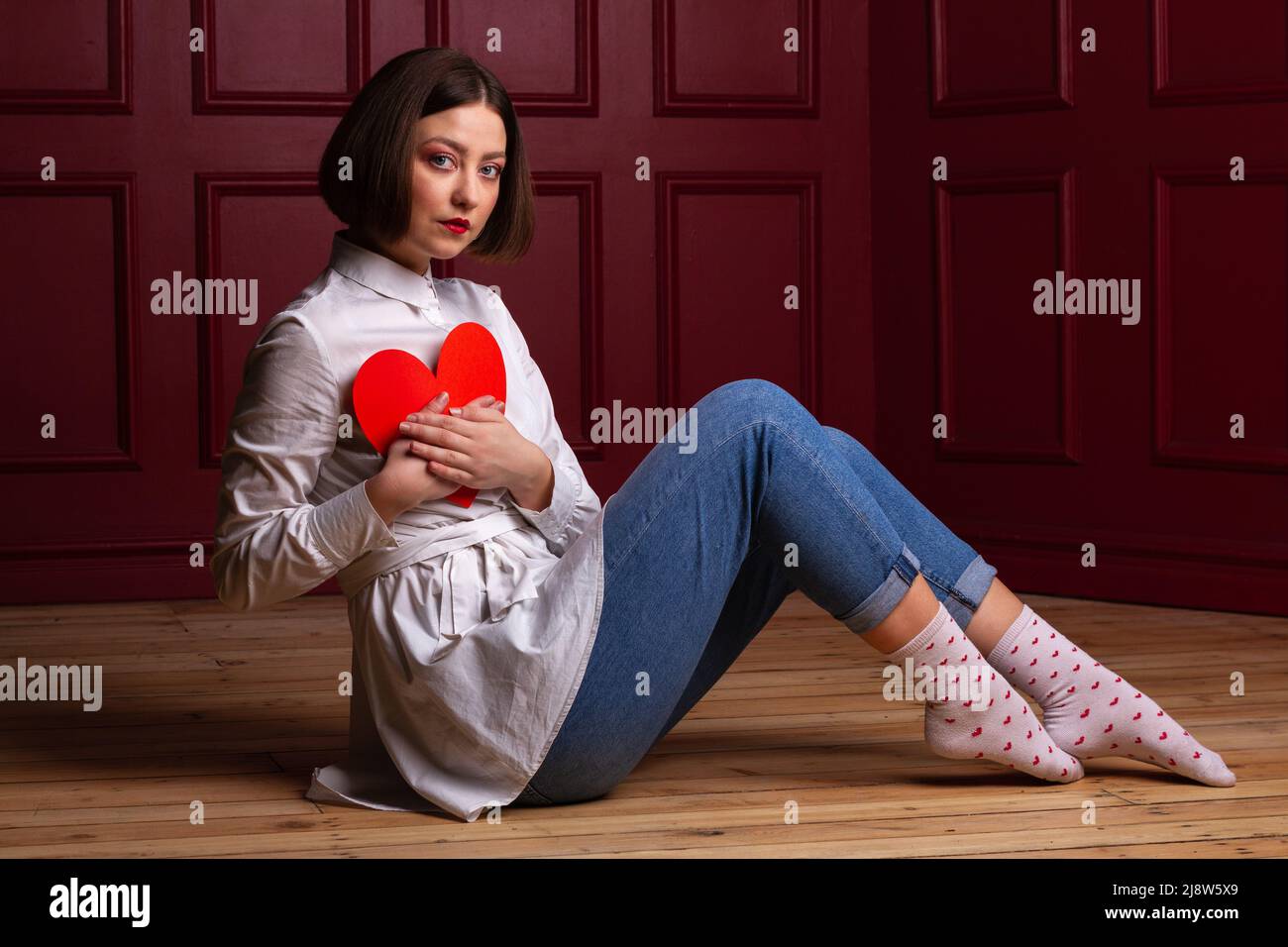 Femme aux cheveux courts assise à côté de l'appareil photo sur un sol en bois et un arrière-plan rouge tenant la forme du cœur rouge sur la poitrine Banque D'Images