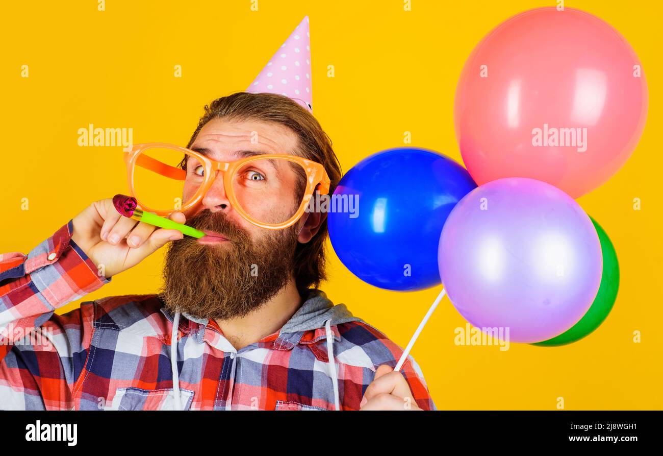 Homme barbu dans un chapeau de fête et des lunettes avec des ballons soufflant de bruit. Fête d'anniversaire. Banque D'Images