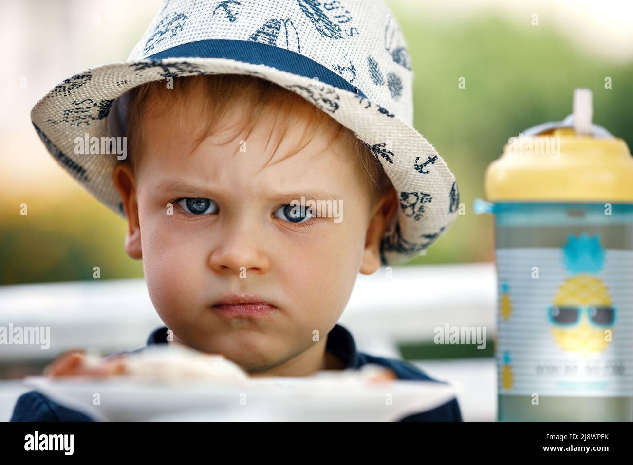 Un petit garçon blond en colère et mécontent de 3 ans ne veut pas manger du pain avec du fromage dans le jardin à la table et de gros plan Banque D'Images