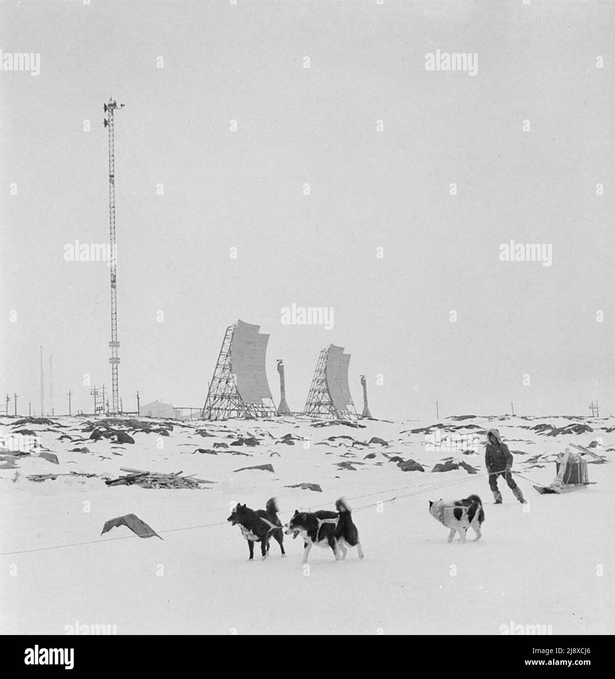 Équipe de chiens et de traîneaux à l'extérieur d'une station radar au Canada. L'image semble être une station Mid-Canada Line (MCL), bien qu'il y ait une petite possibilité il s'agit d'un RADAR AN/FPS-23 de la ligne DEW (FPS-23 généralement utilisé des antennes rectangulaires, pas rond comme dans cette image). La grande tour de gauche est le mât radar, qui dans ce cas a des antennes face à une seule direction, alors que la plupart auraient un deuxième ensemble orienté vers la tour suivante dans la chaîne. Les grandes antennes paraboliques au milieu de l'image font partie du système de diffusion radio troposphérique qui envoie des données au NORAD CA. 1959 Banque D'Images