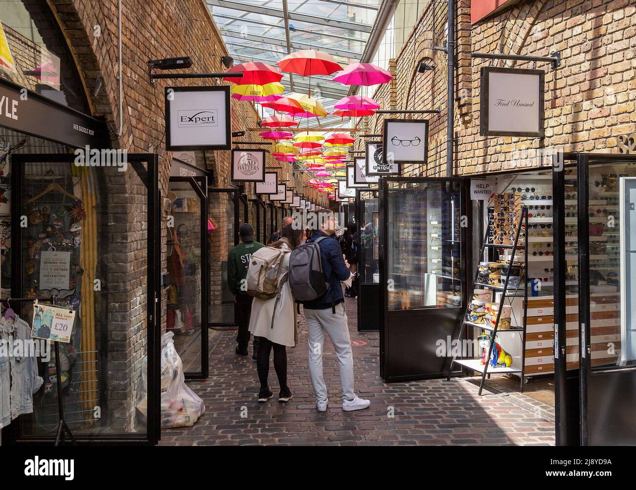 Les clients qui visitent une longue rangée de boutiques de style au marché Camden. Les parasols au-dessus de la passerelle sont indiqués par les mots Camden Market. Banque D'Images