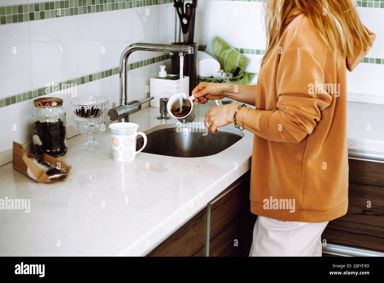 Photo courte d'une jeune femme aux cheveux fins ondulés portant un sweat-shirt marron, en lavant la tasse du café sous l'eau libre. Banque D'Images