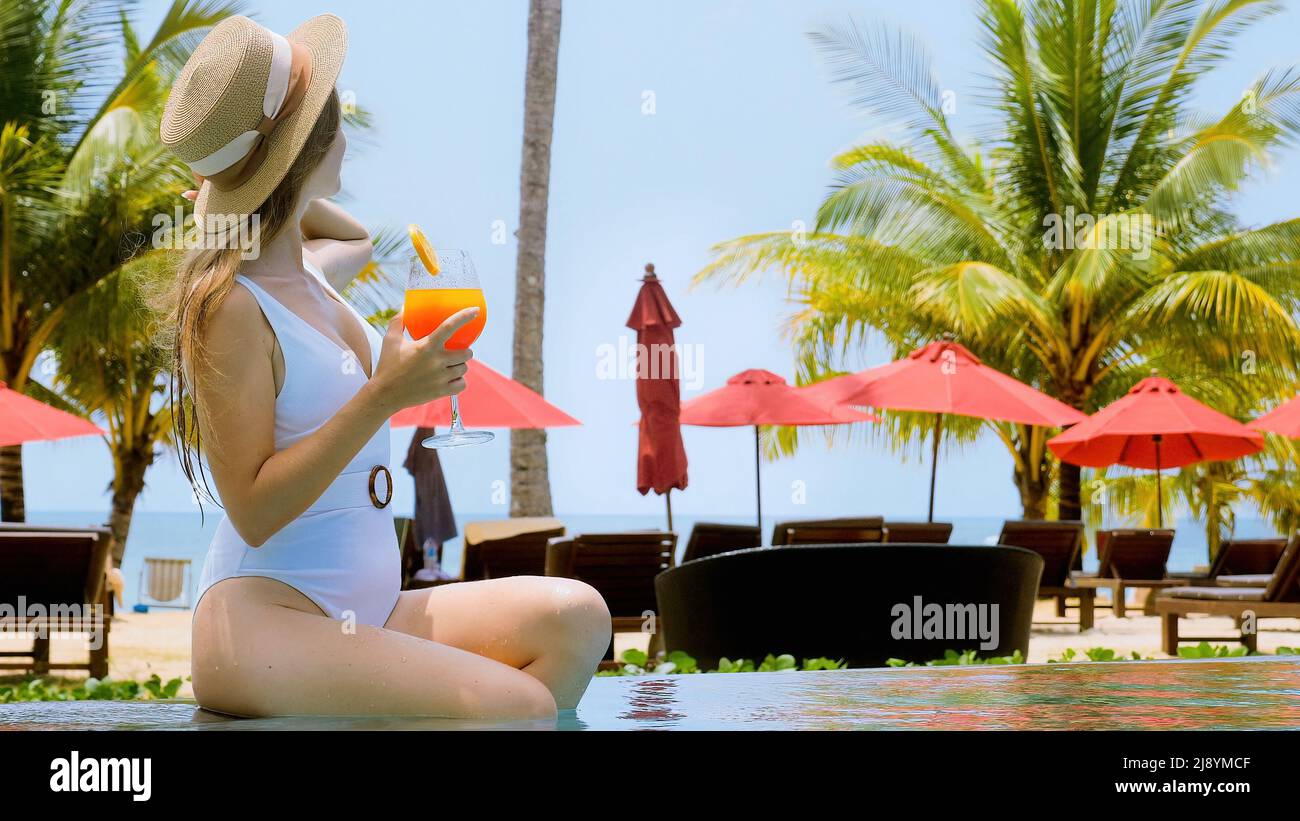 Une jeune femme inconnue au chapeau de paille peut siroter un cocktail dans la piscine extérieure de l'hôtel. Belle fille en maillot de bain blanc relaxant avec boisson de jus Banque D'Images