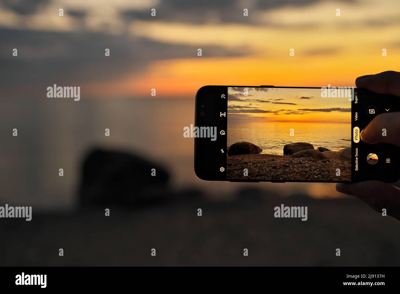 Gros plan d'une main tenant et prenant une photo de smartphone mobile d'une scène de plage au coucher du soleil Banque D'Images