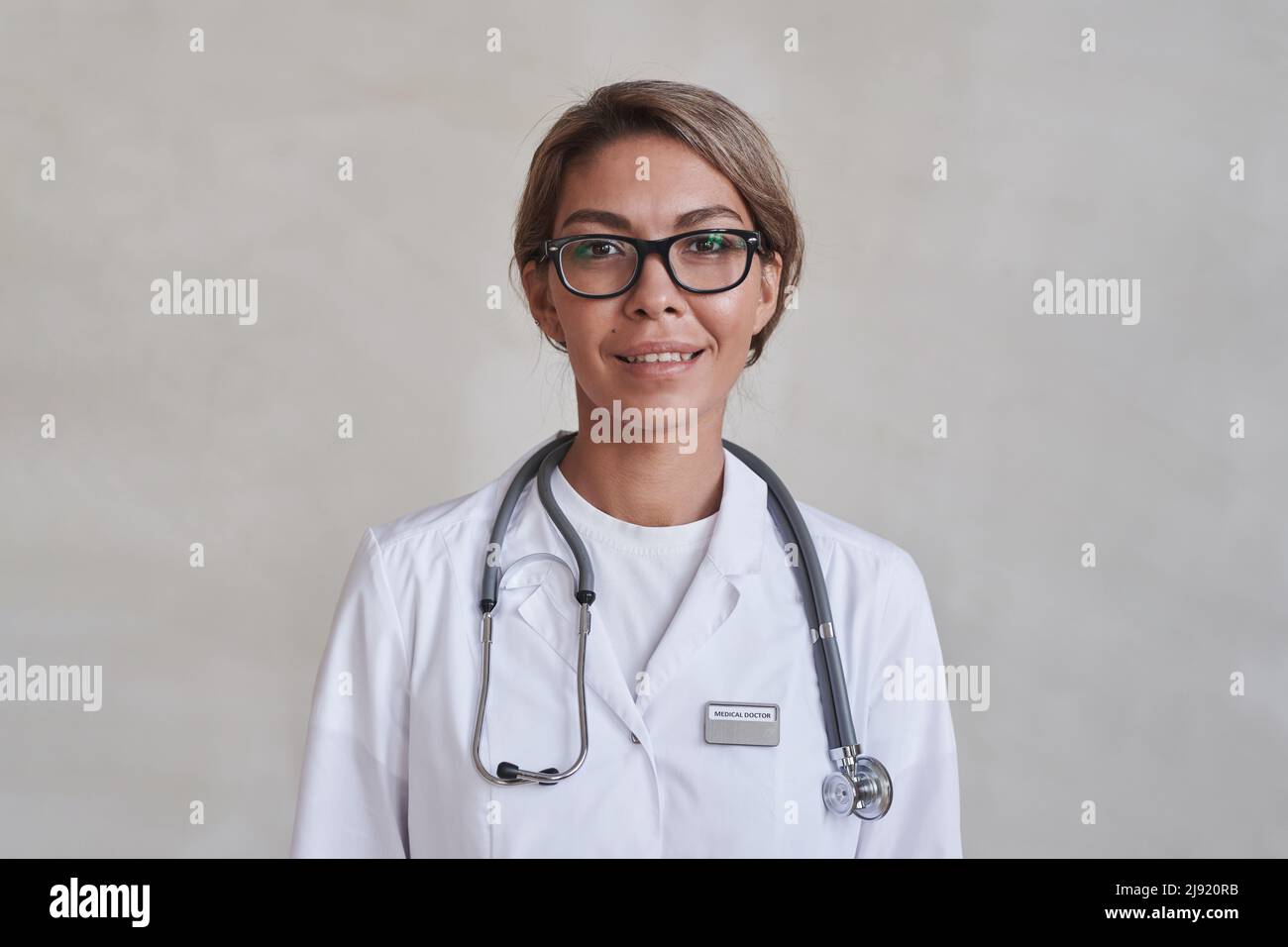 Portrait en gros plan horizontal moyen d'une femme moderne médecin portant des lunettes et un manteau blanc souriant à l'appareil photo Banque D'Images