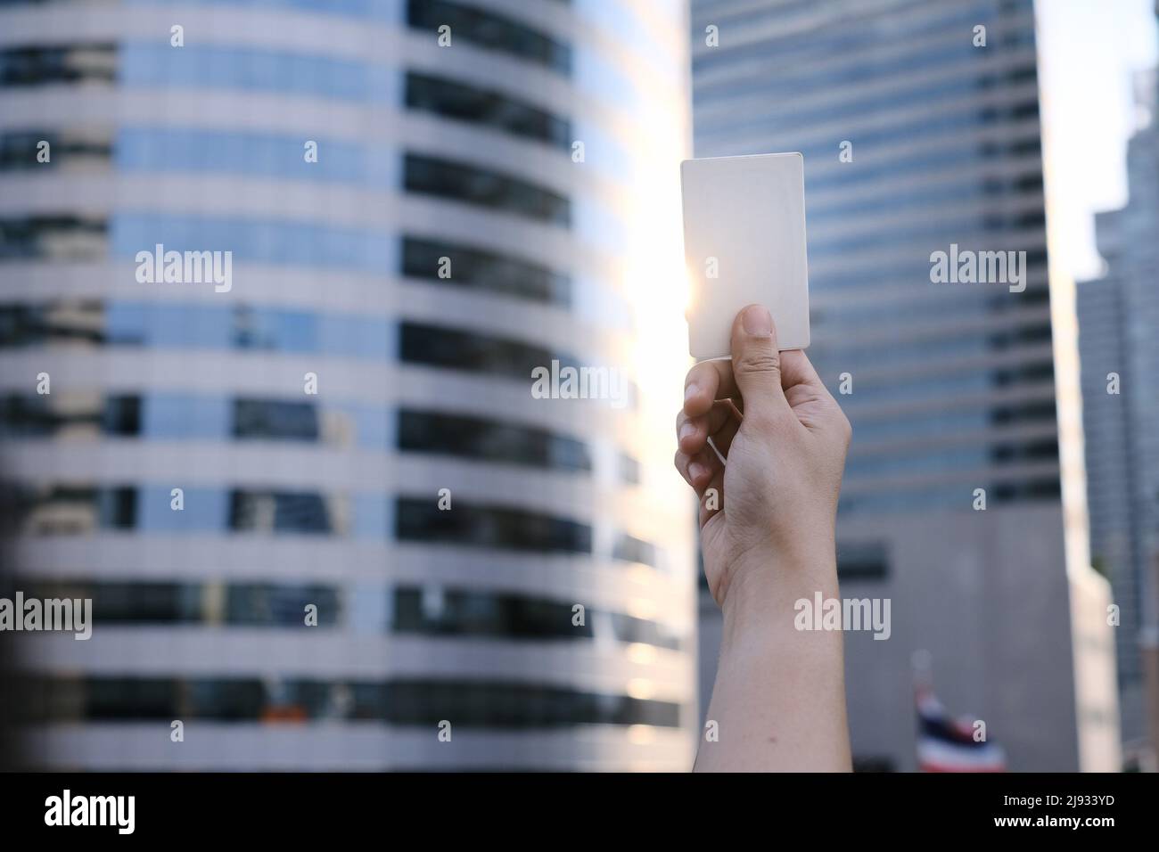 Une main tenant une carte de crédit blanche vierge avec lumière du soleil qui brille à travers sur le côté et les bâtiments élevés en arrière-plan. Banque D'Images