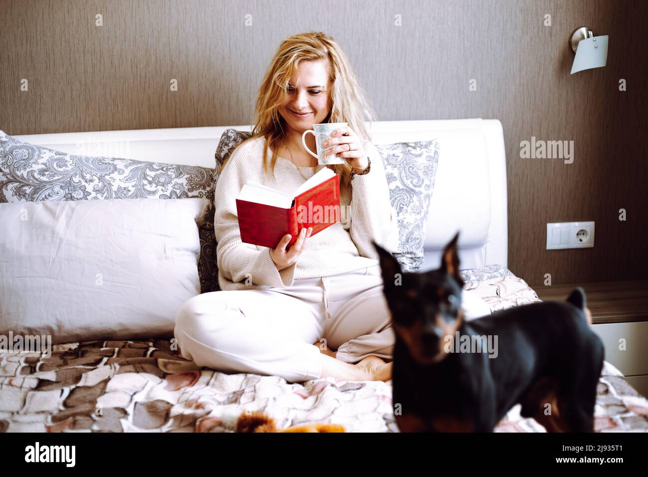 Portrait d'une jeune femme heureuse avec de longs cheveux ondulés équitables portant un chandail beige, un pantalon, assis sur le lit, livre de lecture. Banque D'Images