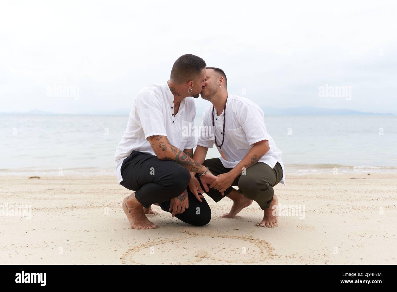 Couple gay embrassant à côté d'un coeur dessiné dans le sable sur une plage tropicale Banque D'Images