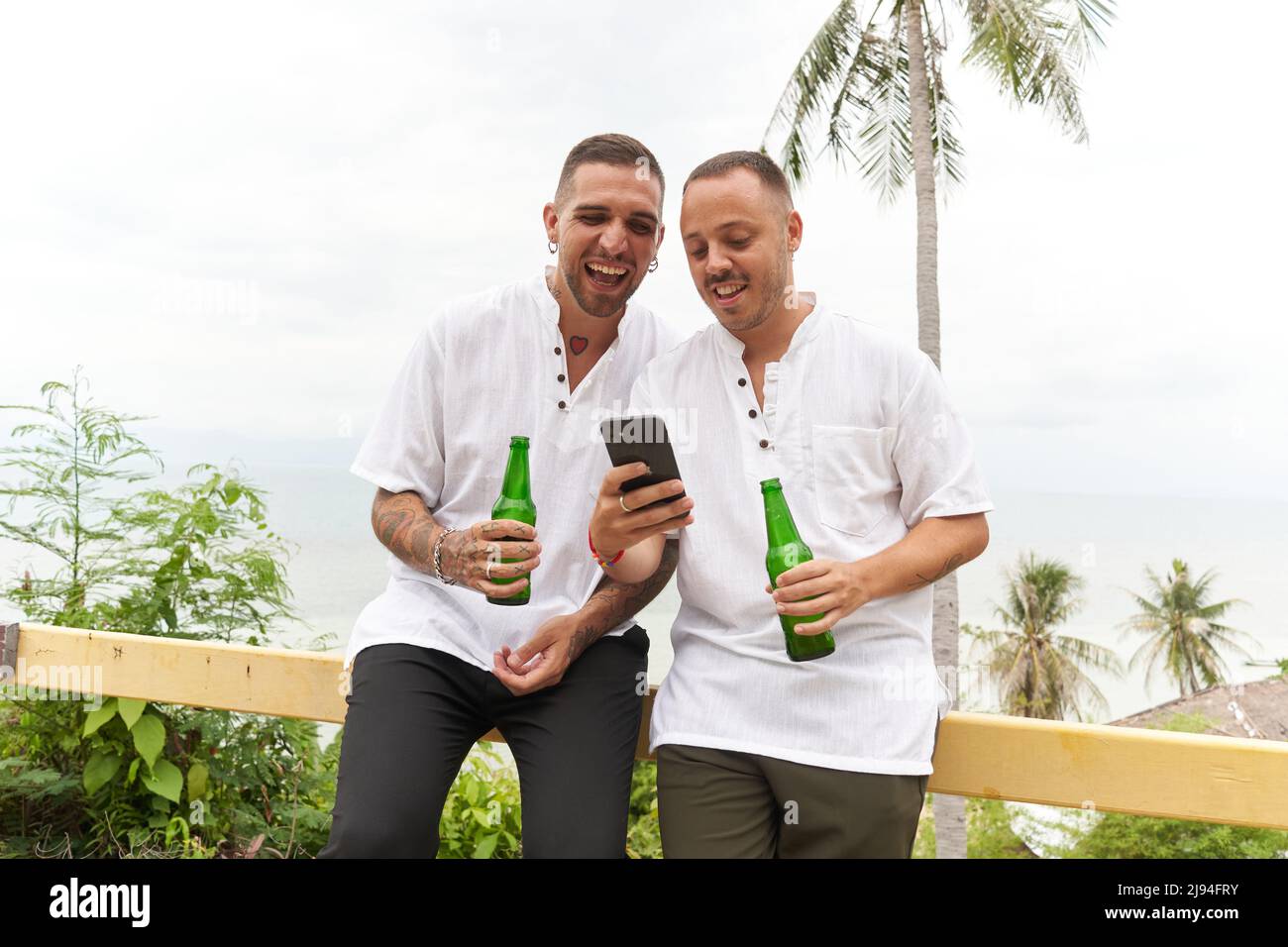 Couple gay utilisant le mobile tout en buvant de la bière dans une terrasse donnant sur la mer Banque D'Images