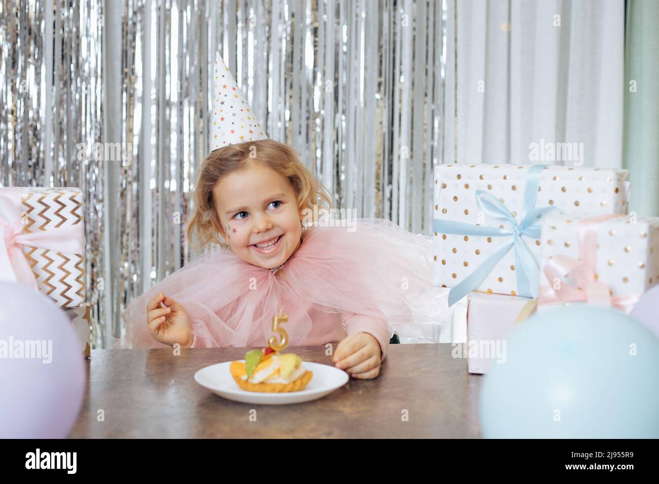 Émotions positives des enfants. Joyeux sourire fille dans la tenue de fête et chapeau de fête est assis devant le gâteau d'anniversaire décoré avec le numéro cinq Banque D'Images