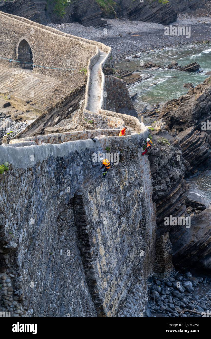Bakio, Espagne - 3 mai 2022 : des techniciens d'accès à la corde et des ouvriers de la construction réparent les murs et le pont en pierre historiques sur la côte basque espagnole Banque D'Images