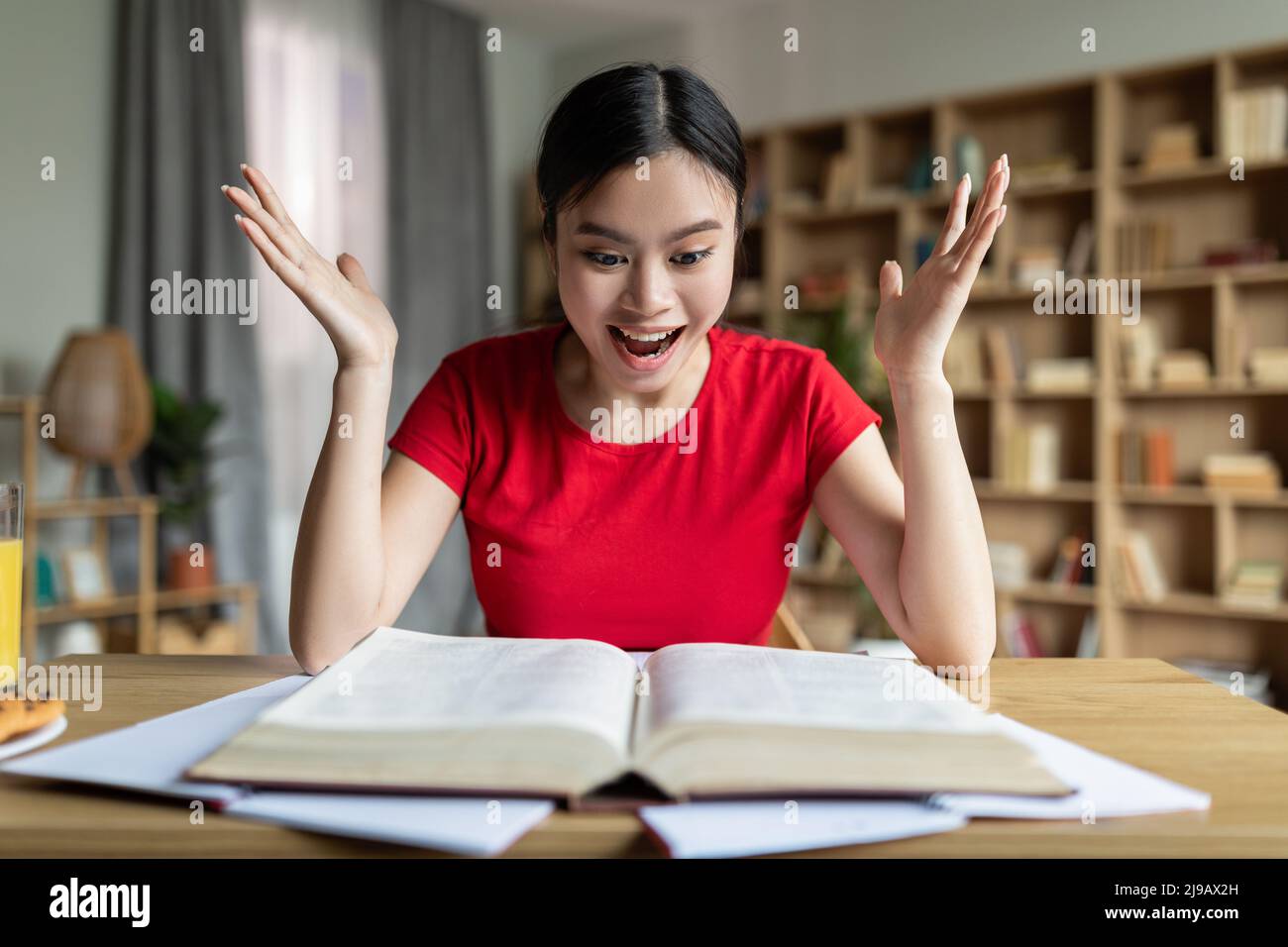 Joyeux affectif intelligent joli jeune asiatique femme étudiant avec la bouche ouverte criant avec joie à la table avec le livre Banque D'Images
