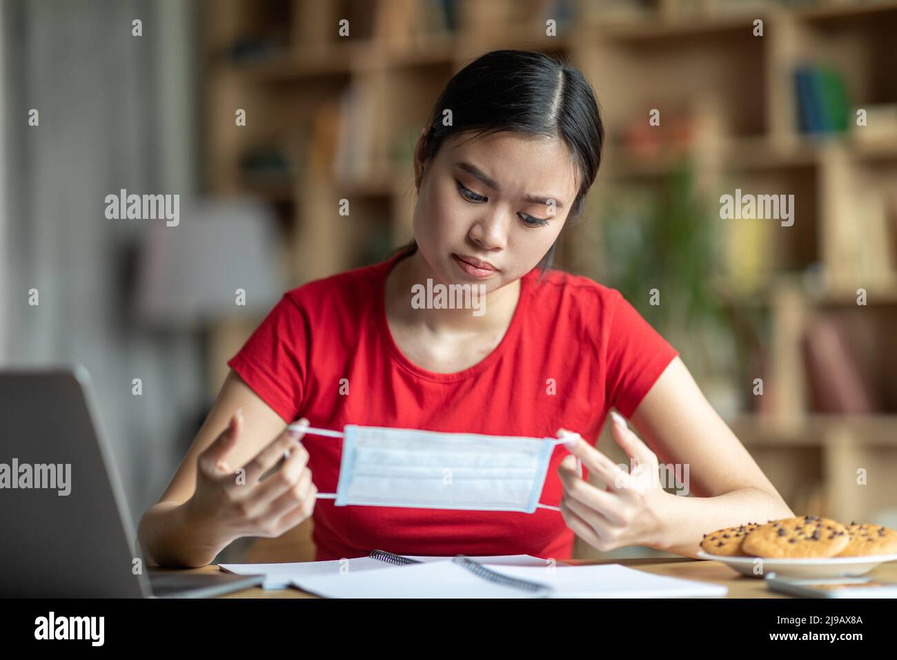 Triste jeune asiatique regarder le masque de protection dans les mains, s'asseoir à la table avec ordinateur portable dans le salon intérieur Banque D'Images