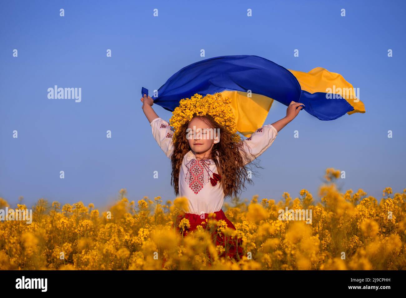 Enfant ukrainien en chemise brodée et couronne jaune dans le champ de fleurs jaunes contre ciel bleu. Drapeau bleu-jaune de l'Ukraine volant dans le vent dans les mains de la petite fille ukrainienne. Priez pour l'Ukraine. Banque D'Images