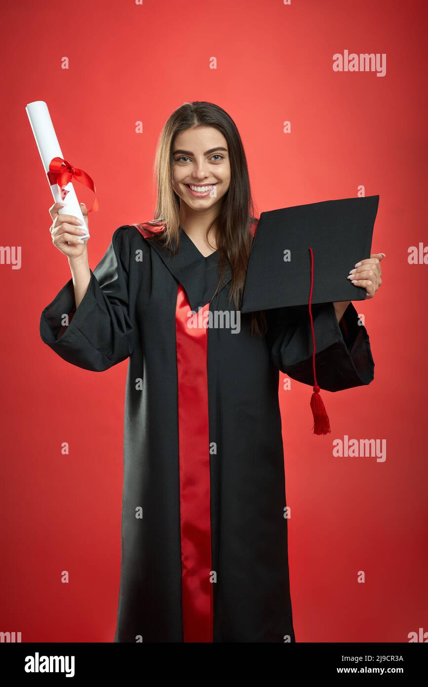 Vue de face d'une jeune fille brunette qui obtient son diplôme d'études collégiales, universitaires et secondaires. Jolie fille regardant l'appareil photo, souriant, tenant diplôme, mortaboard. Isolé sur fond rouge, Banque D'Images Vue de face d'une jeune fille brunette qui obtient son diplôme d'études collégiales, universitaires et secondaires. Jolie fille regardant l'appareil photo, souriant, tenant diplôme, mortaboard. Isolé sur fond rouge, Banque D'Images