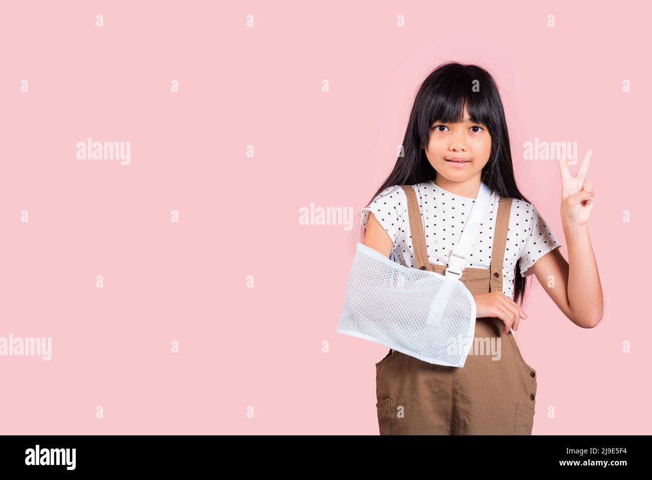 Bras cassé. Asiatique petit enfant de 10 ans os de main cassé de l'accident avec l'attelle de bras montrer le signe V au studio de tir isolé sur fond rose, heureux Banque D'Images