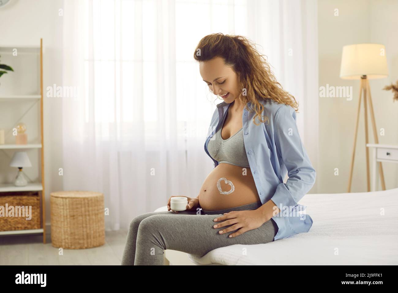 La femme enceinte du neuvième mois s'assoit à la maison sur le lit et sent l'abdomen avec une crème anti-stretch. Banque D'Images