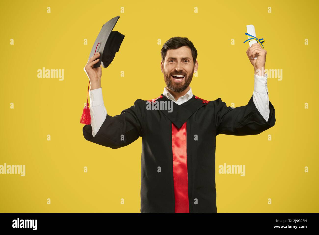 Vue de face d'un diplôme d'études et d'un mortarboard heureux. Maître, Bachelor debout, souriant, regardant la caméra, portant une robe de troisième cycle, joyeux. Isolé sur fond jaune. Banque D'Images