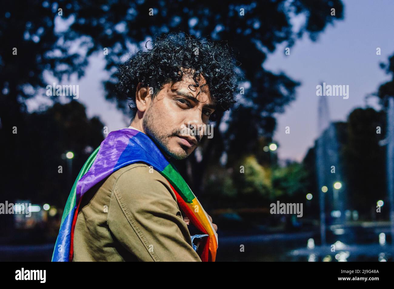 portrait d'un homme à l'extérieur avec des curlers, avec un drapeau de fierté gay sur ses épaules, regardant l'appareil photo. Banque D'Images