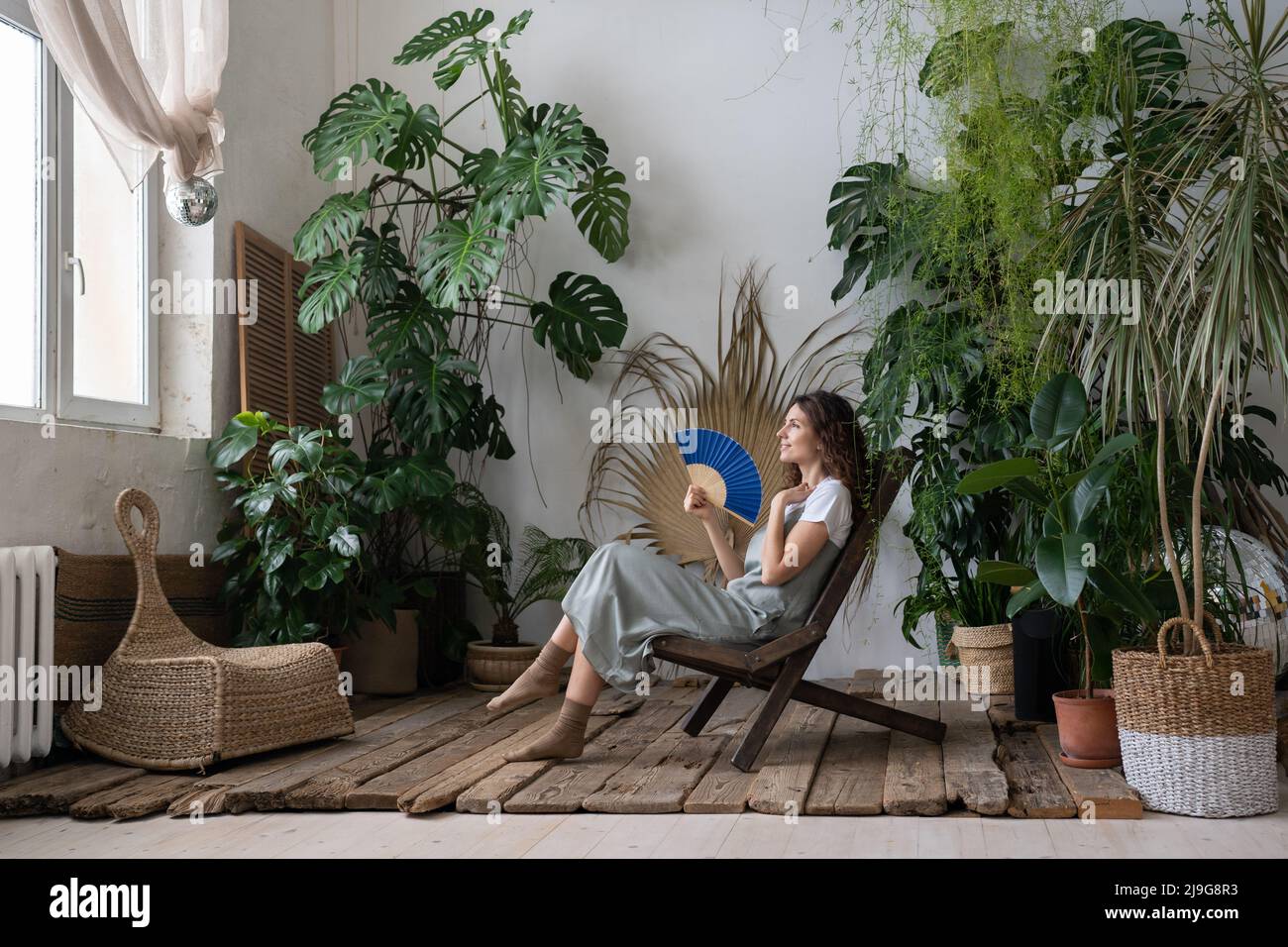 Jeune femme rêveuse et détendue utilisant un ventilateur de papier tout en se relaxant dans le beau jardin rafraîchissant de la maison Banque D'Images