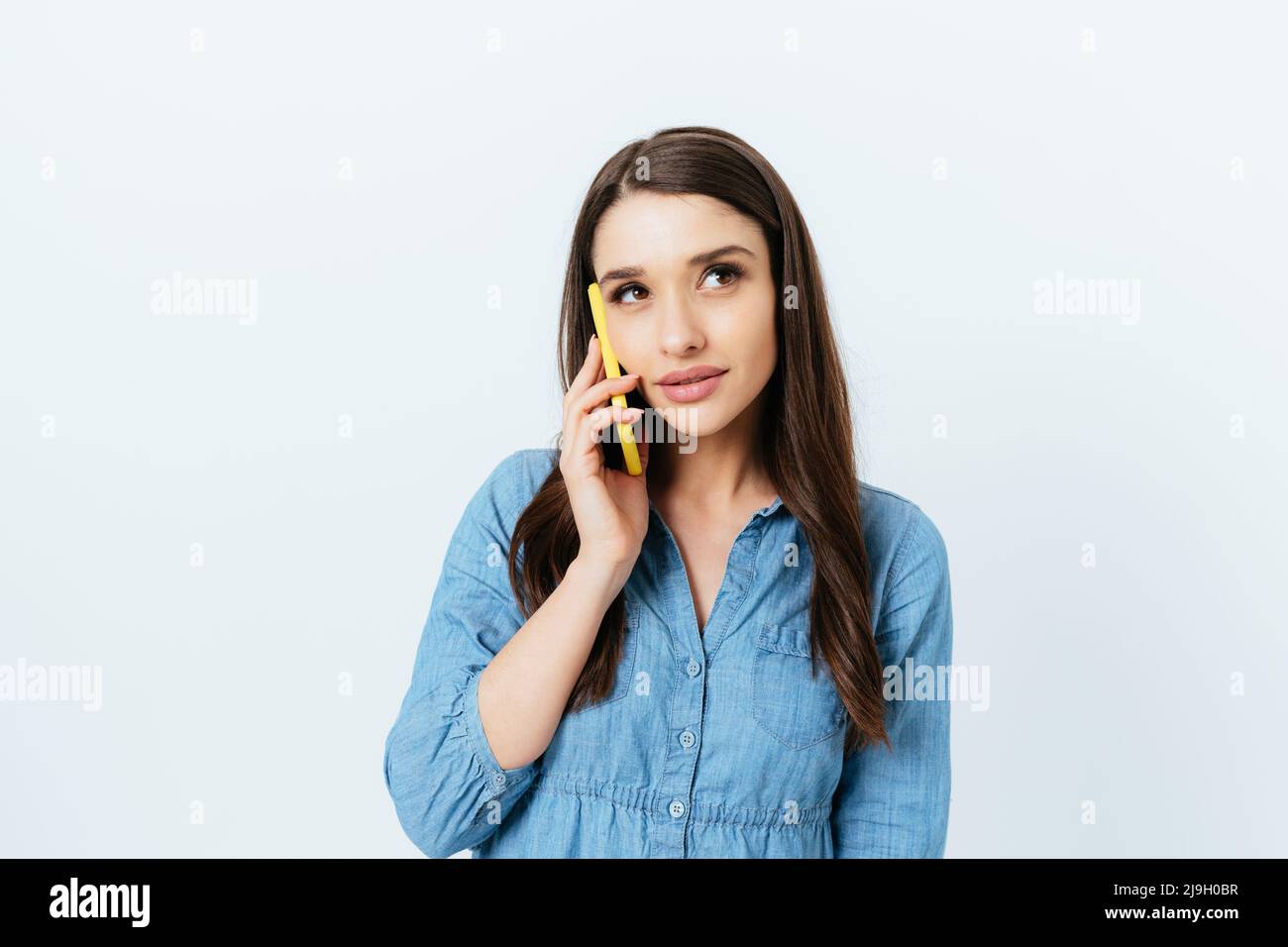 Élégante jeune femme brunette parlant sur téléphone mobile souriant légèrement et regardant loin, fond blanc studio Banque D'Images