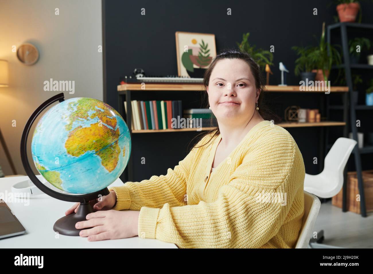 Portrait horizontal moyen de jeune fille joyeuse avec le syndrome de Down assis au bureau avec le globe sur lui regardant l'appareil-photo Banque D'Images