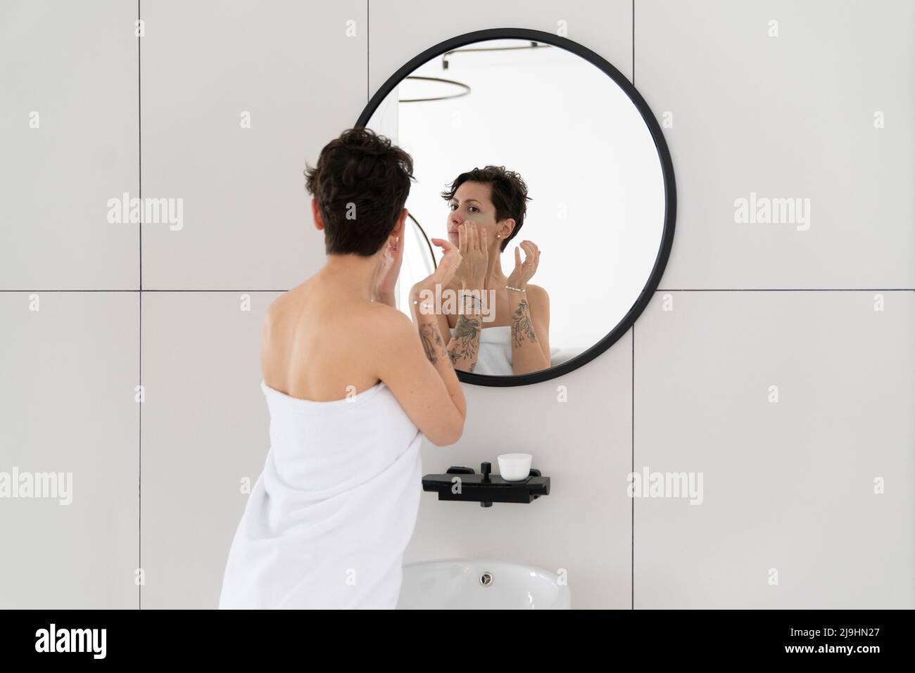 Femme enveloppée dans une serviette appliquant un masque oculaire regardant le miroir Banque D'Images