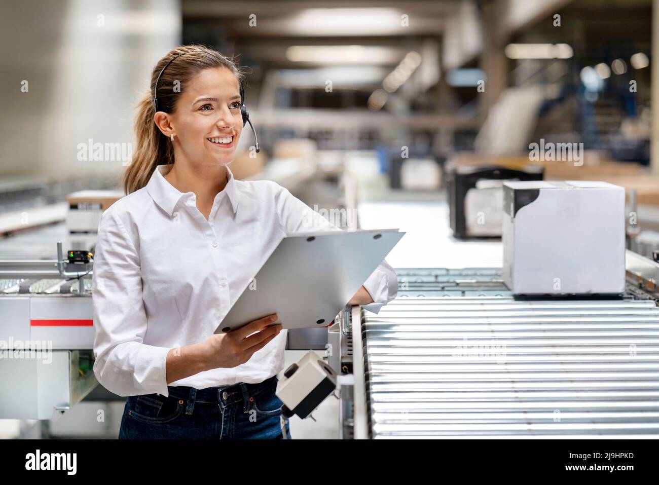 Une femme d'affaires heureuse avec un presse-papiers portant un micro-casque en usine Banque D'Images