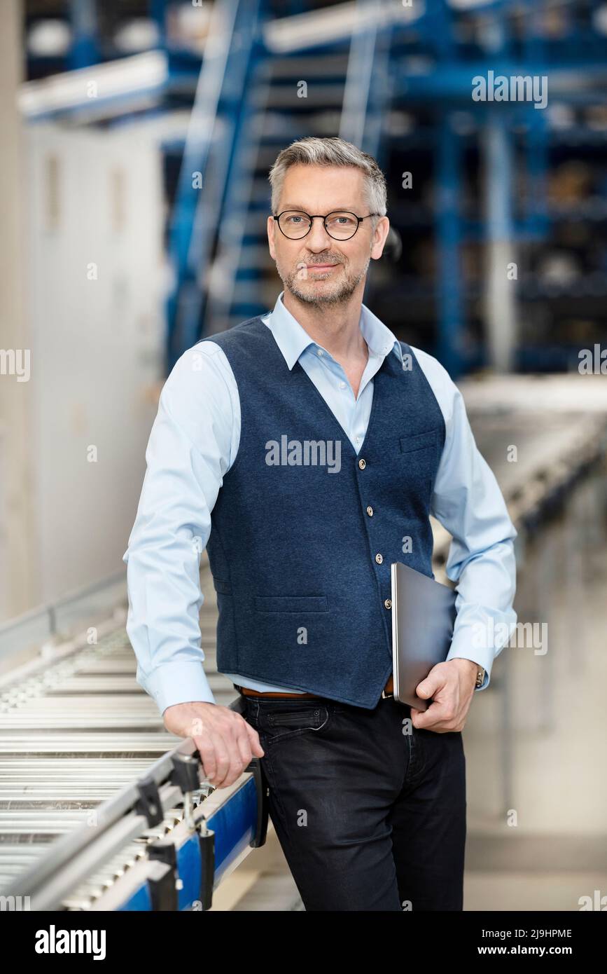 Homme d'affaires confiant avec un ordinateur portable par tapis roulant en usine Banque D'Images