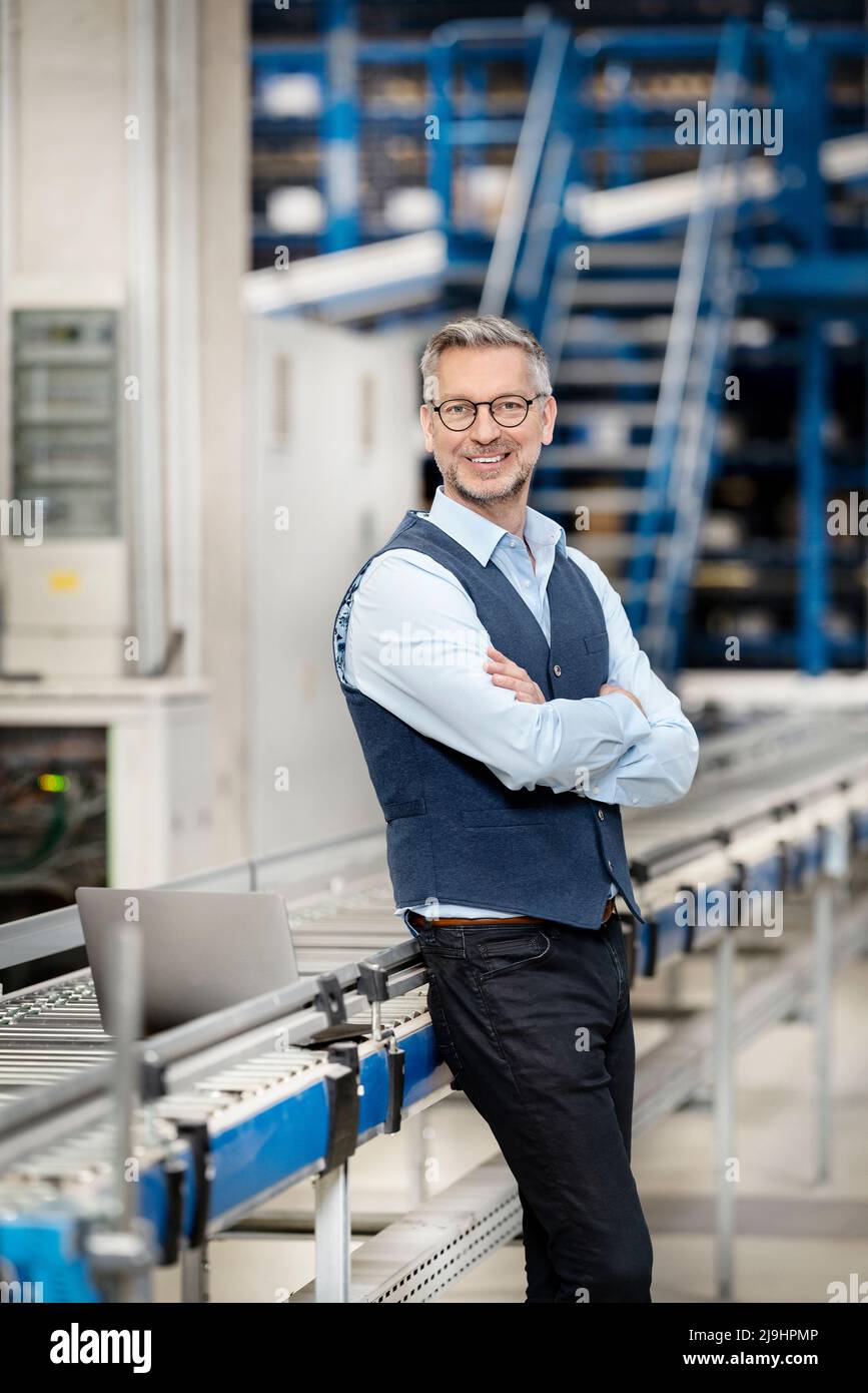 Homme d'affaires heureux avec les bras croisés par le tapis de convoyeur à l'usine Banque D'Images