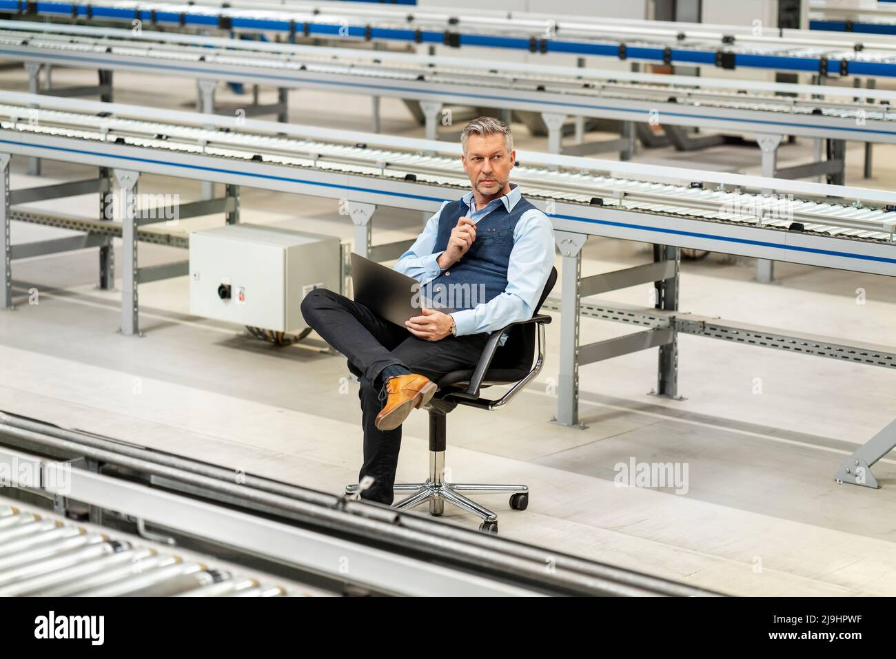 Homme d'affaires avec ordinateur portable assis au milieu d'un tapis roulant à l'usine Banque D'Images