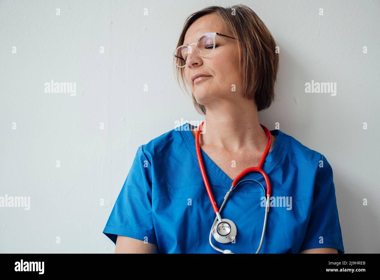 Femme médecin exténuée avec les yeux fermés penchée sur le mur blanc reposant à l'hôpital Banque D'Images