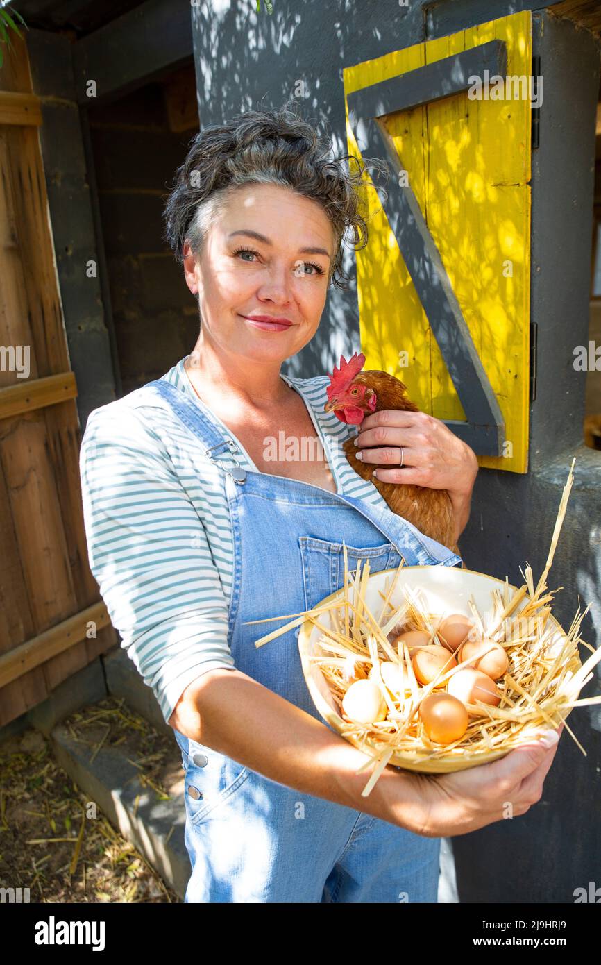 Femme mûre souriante avec une poule et un bol d'œufs debout devant le coop de poulet Banque D'Images