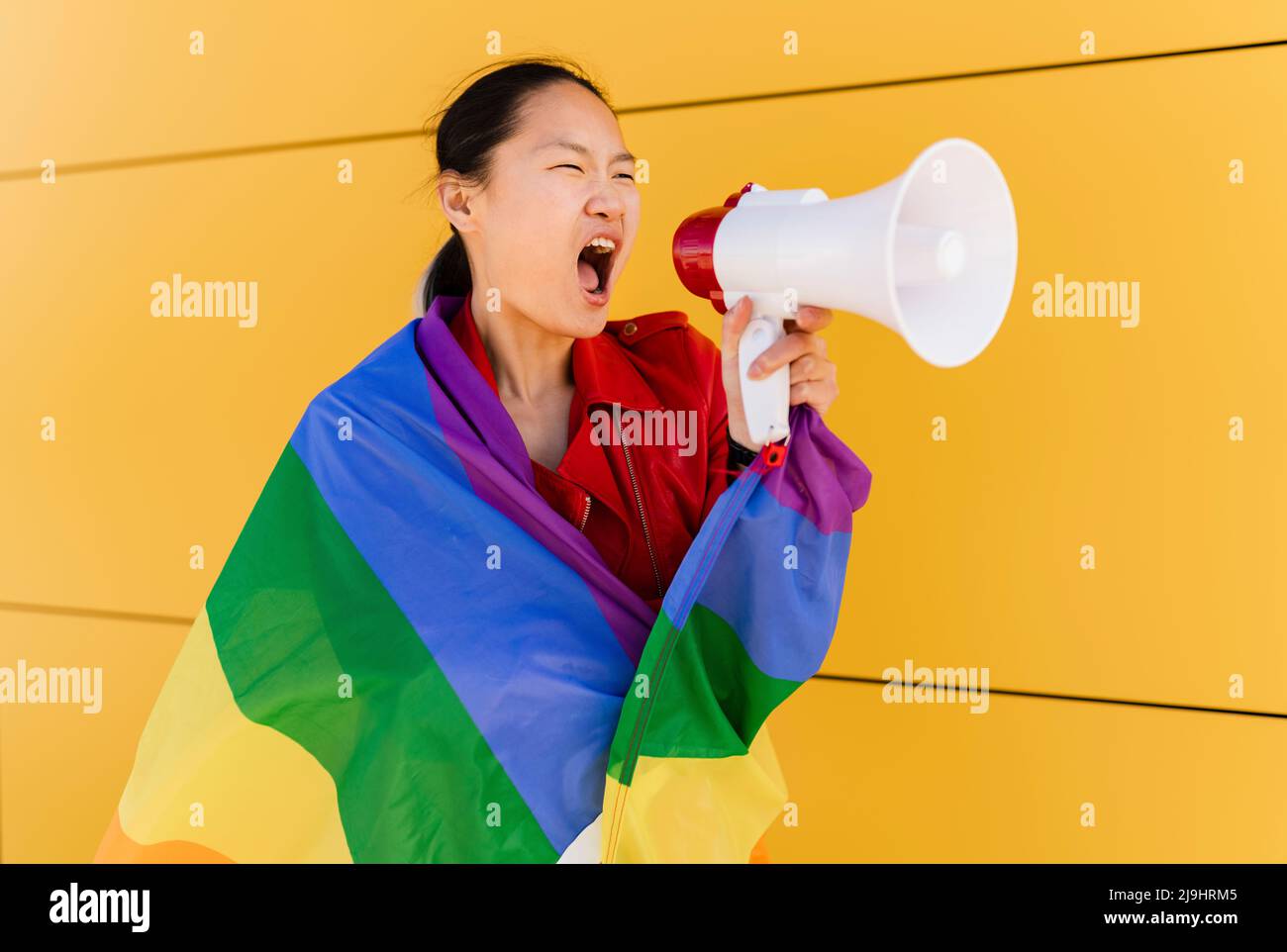 Femme enveloppée dans un drapeau arc-en-ciel criant à travers le mégaphone par le mur jaune Banque D'Images