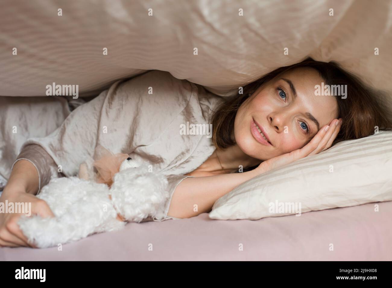Femme mûre souriante avec jouet ours en peluche couché sous une couverture sur le lit à la maison Banque D'Images