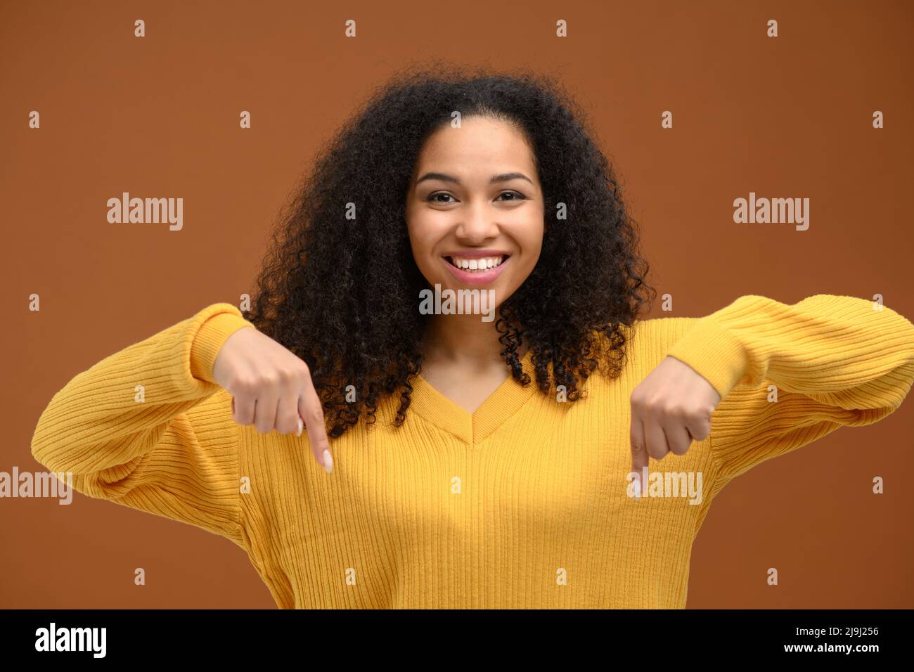 Charmante jeune femme afro-américaine pointe l'index vers le bas, la promotion de la nouveauté isolée sur fond marron, concept publicitaire Banque D'Images