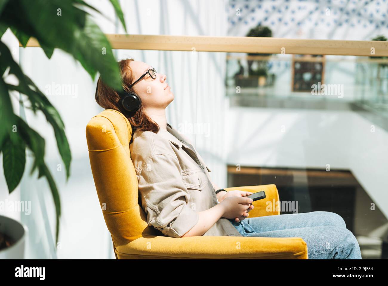 Jeune brune adolescente jeune fille collégiale ou lycéenne dans des lunettes à l'aide d'un téléphone mobile, écouter de la musique dans un casque dans une chaise jaune dans un endroit public Banque D'Images