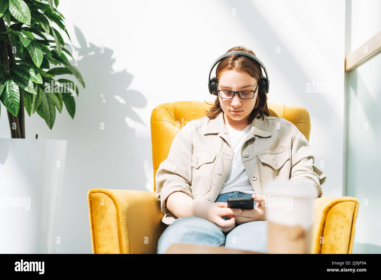 Jeune brune adolescente jeune fille collégiale ou lycéenne dans des lunettes à l'aide d'un téléphone mobile, écouter de la musique dans un casque dans une chaise jaune dans un endroit public Banque D'Images